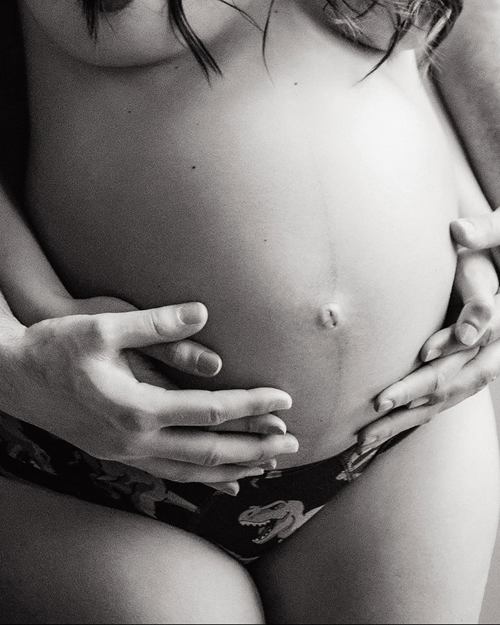 Close-up black-and-white maternity portrait of an expecting couple holding their baby bump during a maternity boudoir session.